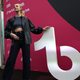 A woman in a black shirt, coat and pants holds up the TikTok logo to her right.